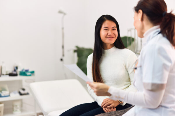 A smiling Asian female patient talking with a female doctor, sitting in the medical office. A content happy adult Asian patient visited a female doctor, talking about her blood results, looking healthy.